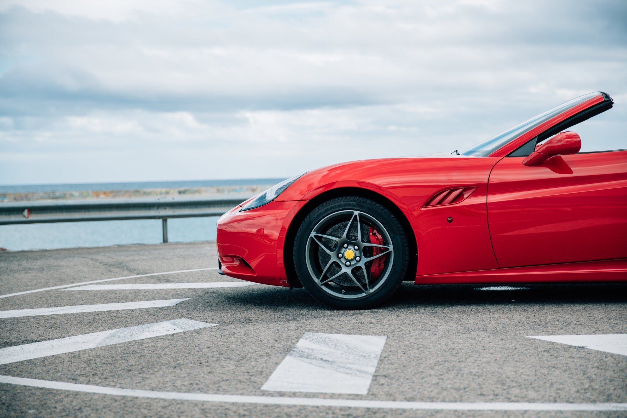 Red convertible sports car parked on a coastal road with cloudy sky in the background.