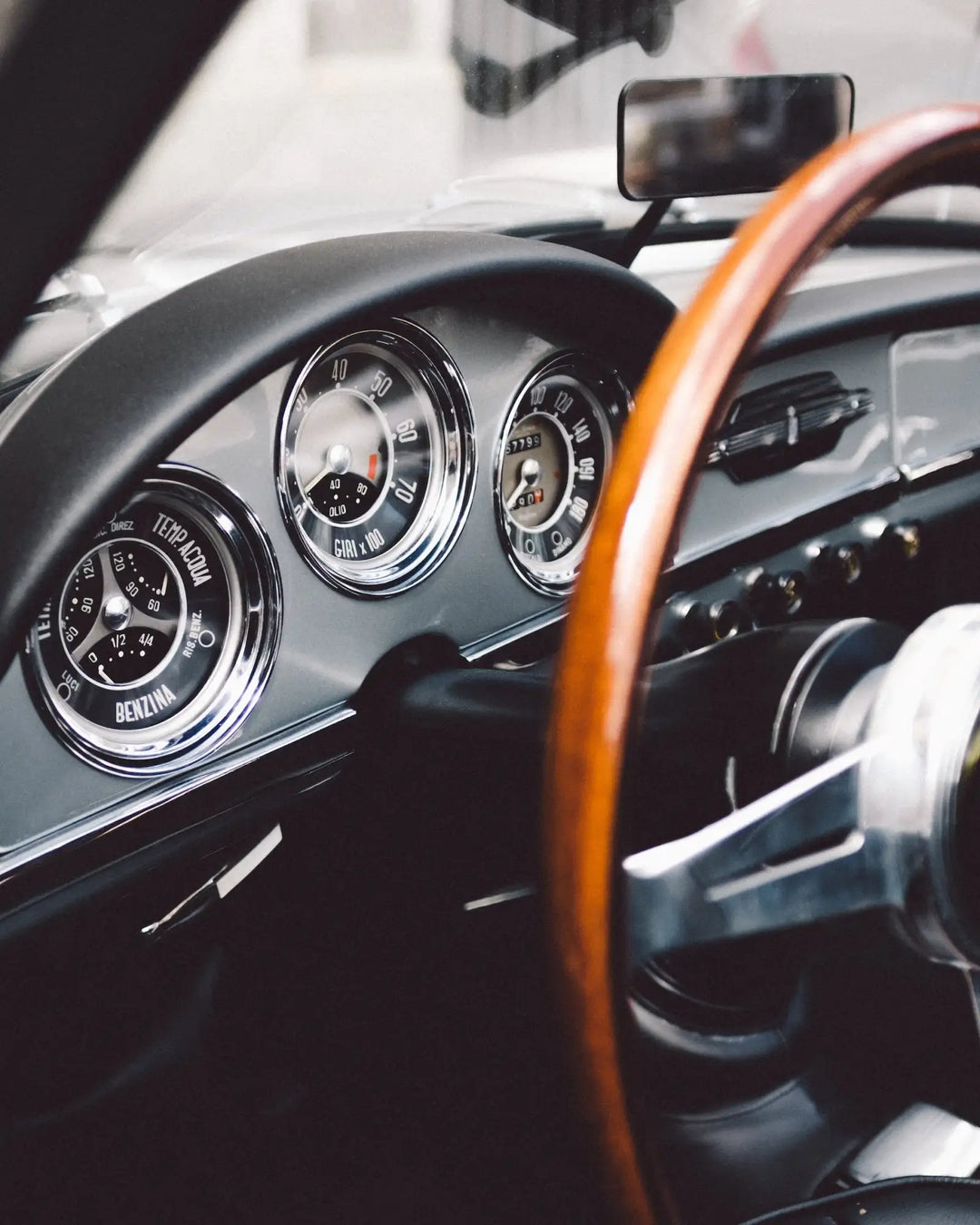 The image shows a close-up of the dashboard and steering wheel from an old, classic car with chrome-rimmed analog gauges set into a dark gray panel.