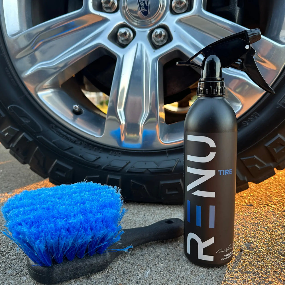TIRE cleaning spray bottle and blue brush in front of a rugged tire with shiny chrome wheel on pavement